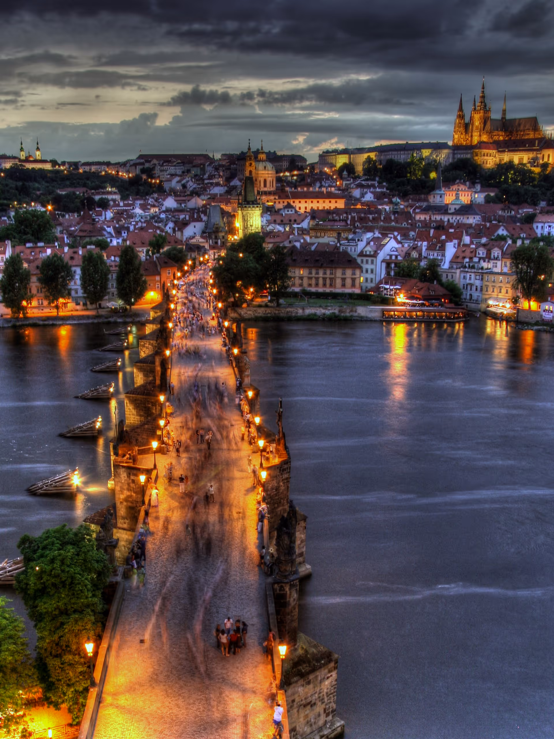 Illuminated Charles Bridge with people walking over the Vltava River at dusk in Prague, with Prague Castle visible in the background under a cloudy sky.