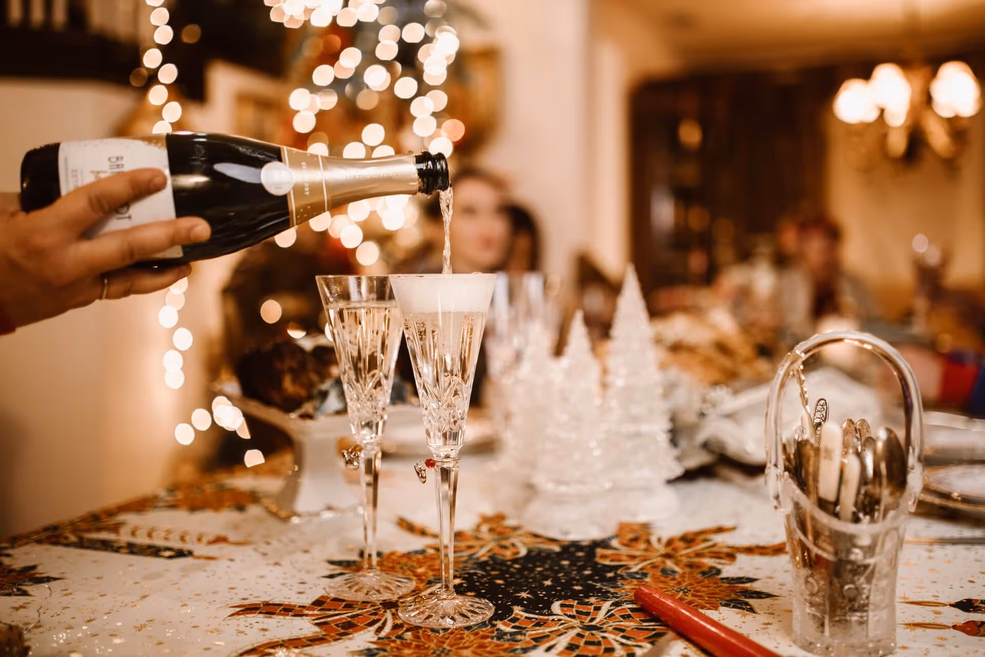 Hand pouring champagne into crystal flutes on a festive table with holiday decorations and blurred background lights.