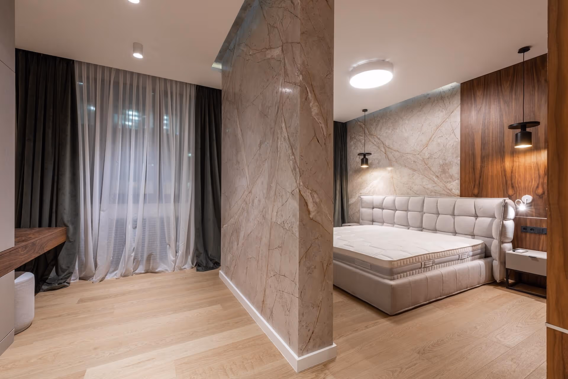 Modern bedroom with light wood flooring, a large bed with tufted headboard, marble accent walls, and sheer white curtains over a window.