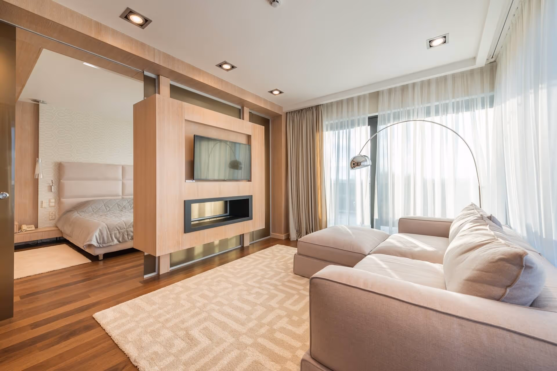Modern living room with a beige sectional sofa, floor lamp, large window with sheer curtains, and a wooden partition wall with a mounted TV and fireplace, adjacent to a bedroom.