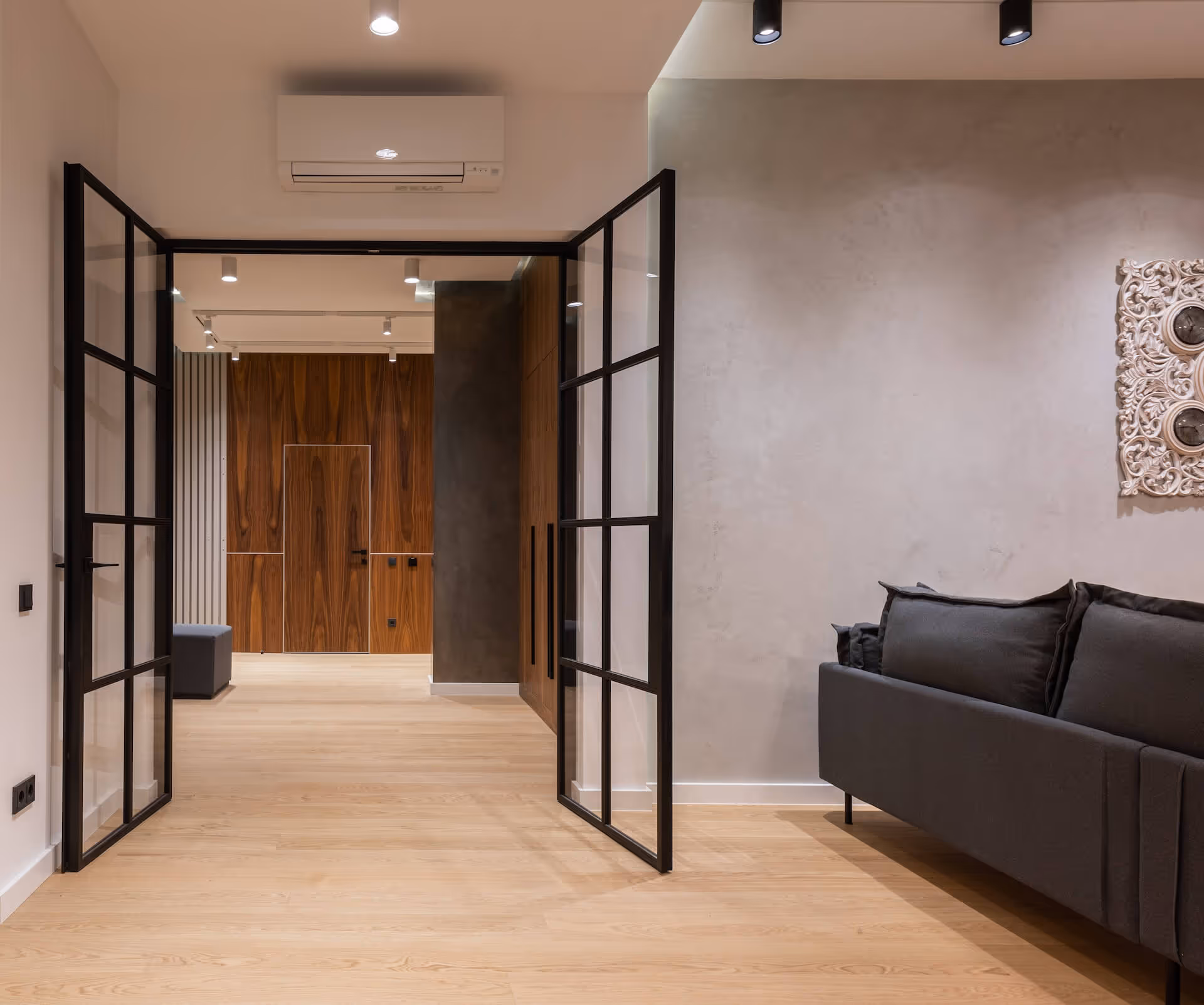 Modern interior with open black-framed glass doors, light wood flooring, dark gray sofa, and a wall clock with ornate white frame.