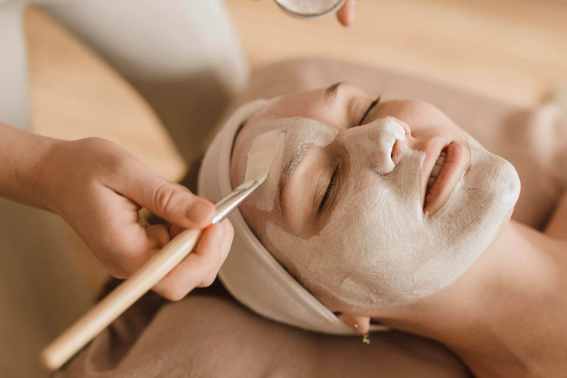 Person with eyes closed receiving a facial mask treatment applied with a brush.